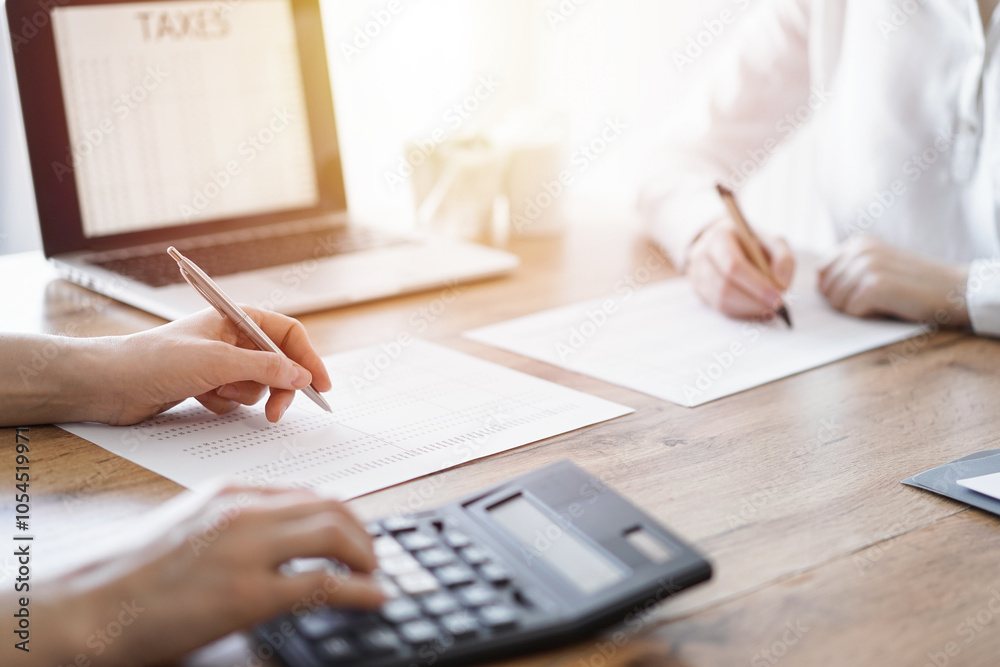 Two accountants using a laptop computer and calculator while counting taxes at wooden desk in office. Teamwork in business audit and finance Two accountants using a laptop computer and calculator while counting taxes at wooden desk in office. Teamwork in business audit and finance