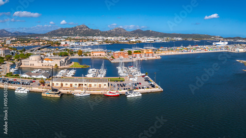 Aerial view of Olbia, Sardinia, showcasing a marina with yachts, a waterfront promenade, and a mix of modern and traditional architecture along the coast.