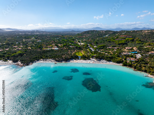 Aerial view of a crescent shaped beach in Sardinia, Italy, with turquoise waters, lush greenery, scattered buildings, and hills in the background.