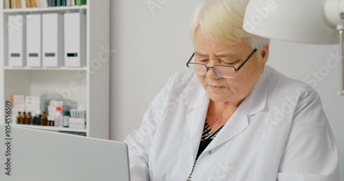 Senior woman doctor hands using laptop computer and working at desk in hospital office. Female heathcare assistant keeps medical records on her laptop. photo