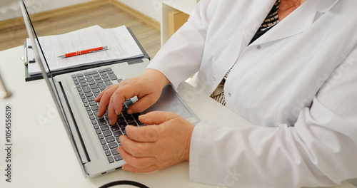 Side view of senior woman doctor hands using laptop computer and working at desk in hospital office. Female heathcare assistant keeps medical records on her laptop.