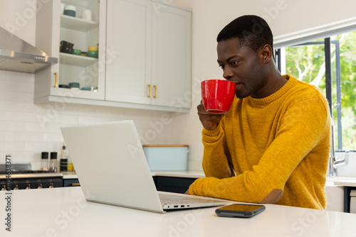 African american man in kitchen drinking coffee while using laptop, at home