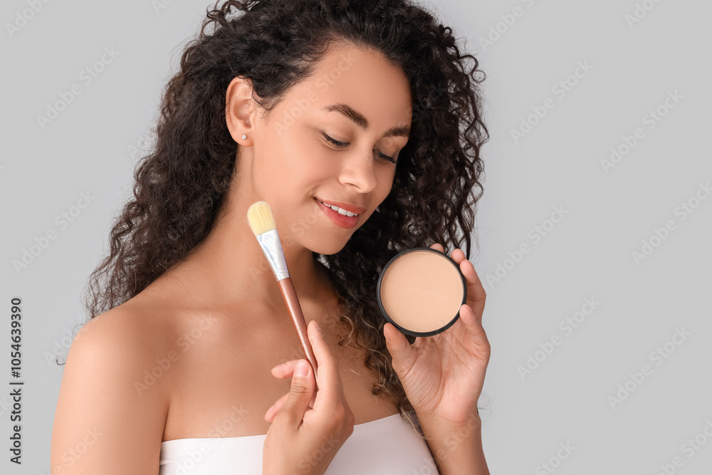 Beautiful young happy African-American woman with makeup brush and powder on grey background Beautiful young happy African-American woman with makeup brush and powder on grey background