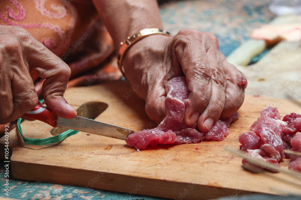 A close-up photo showing the hands of an elderly woman cutting raw red meat using a small knife on a wooden chopping board A close-up photo showing the hands of an elderly woman cutting raw red meat using a small knife on a wooden chopping board
