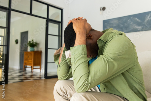 African american man sitting on couch at home, holding head, feeling stressed during holidays photo