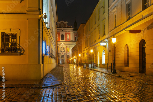 A narrow cobblestone street in Poznan, Poland, illuminated by warm streetlights at night. The scene features historic buildings and the facade of the Fara Church in the background