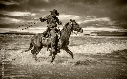 A sepia toned mage of a cowboy riding a horse in the water causing splashes at a beach with a dramatic sky The image is from Rosarito Mexico