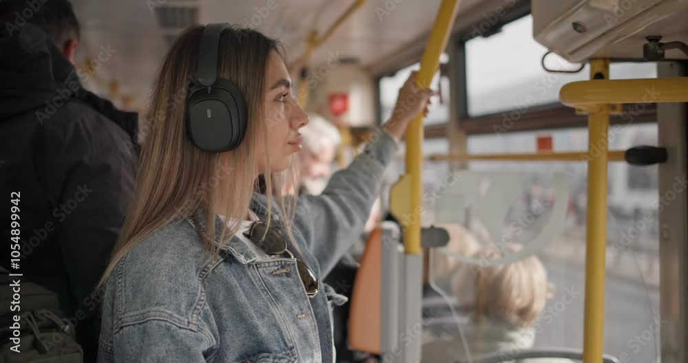 Handheld video of a young girl in a casual denim jacket and wireless headphones riding a city tram, holding onto a handrail, watching and listening to music Handheld video of a young girl in a casual denim jacket and wireless headphones riding a city tram, holding onto a handrail, watching and listening to music