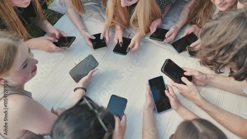Group of teenagers lying on grass using smartphones together around white table.