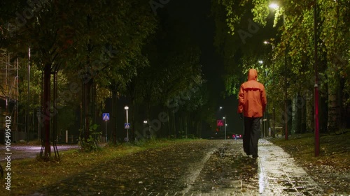 Man in Orange Raincoat Walking Down Wet Street in Strong Rain at Night, Walking Away from Camera, Storm and Bad Weather with Trees Blowing in the Wind photo