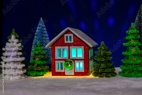 Red house with turquoise light in the windows decorated with a Christmas wreath among green fir trees