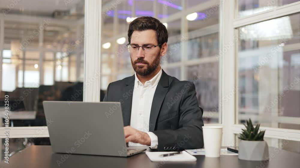 Satisfied businessman in a suit sits at his desk in an office and takes his glasses off after working on his laptop. Manager bank lawyer employee takes a deep breath and looks around the office. Satisfied businessman in a suit sits at his desk in an office and takes his glasses off after working on his laptop. Manager bank lawyer employee takes a deep breath and looks around the office.