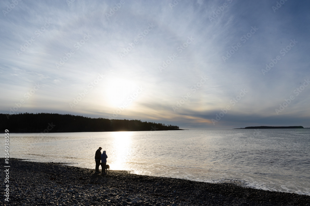 Fototapeta premium Beautiful sunrise or sunset at a beach with a corona around the sun showing an elderly retired couple with their dog in silhouette standing on the rocks looking out watching the water of the ocean