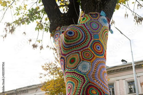 Artistic crochet wrapped around a tree trunk in vibrant colors. Suitable for concepts of creativity, street decor, and unique city installations.