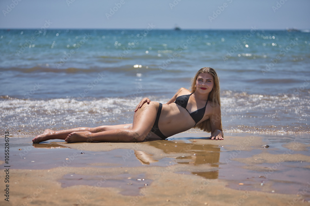 Young, beautiful, blue-eyed, blonde woman in a black bikini posing lying on the shore of the beach with the sea in the background. Concept beauty, fashion, trend, femininity. Young, beautiful, blue-eyed, blonde woman in a black bikini posing lying on the shore of the beach with the sea in the background. Concept beauty, fashion, trend, femininity.