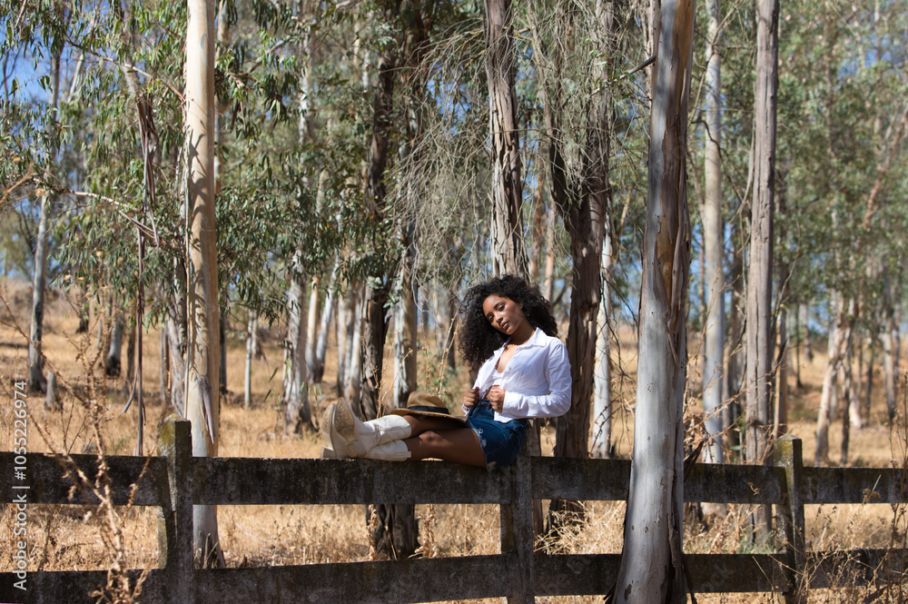 Fototapeta premium woman, young, beautiful, Latin, brunette with curly hair dressed in white shirt, denim skirt, white boots and hat, sitting with her legs stretched out on the forest fence. The woman rests.