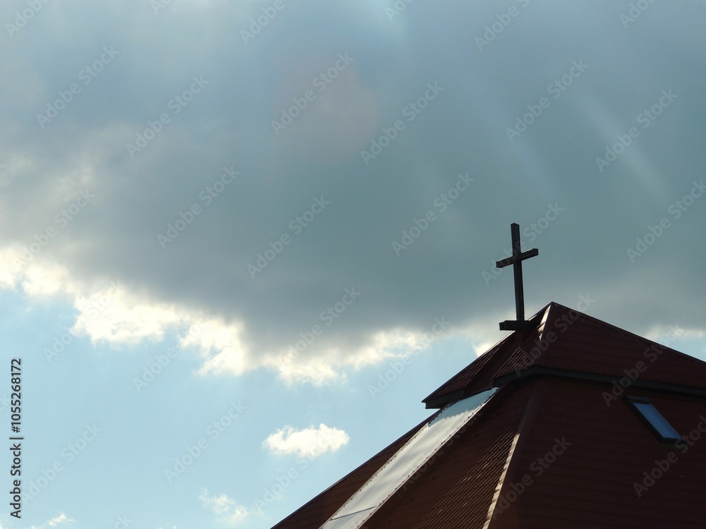 The tiled roof of a Protestant church with a cross on top against a blue sky with a large gray cloud in the background The tiled roof of a Protestant church with a cross on top against a blue sky with a large gray cloud in the background