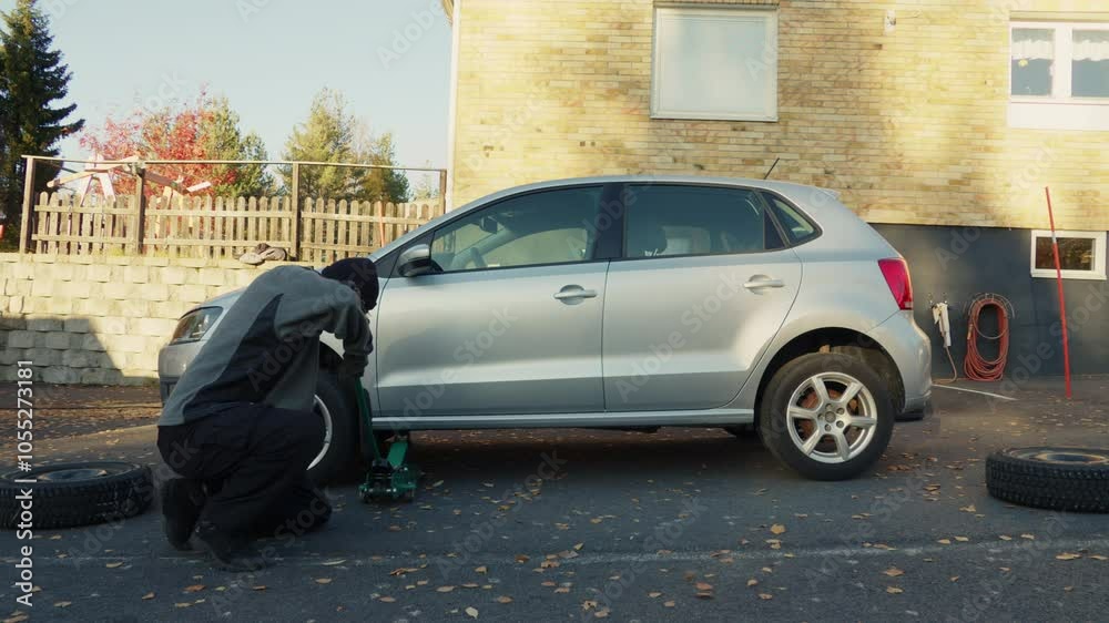 Auto Mechanic Man Using Hydraulic Car Jack to Lift and Raise Silver Car for Service and Changing Tyres, Vehicle Repair in Driveway Outside Garage on Sunny Autumn Day Auto Mechanic Man Using Hydraulic Car Jack to Lift and Raise Silver Car for Service and Changing Tyres, Vehicle Repair in Driveway Outside Garage on Sunny Autumn Day