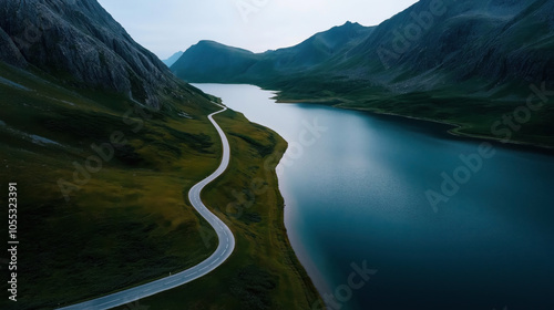 Fototapeta Naklejka Na Ścianę i Meble -  Aerial view of a winding road alongside a serene lake surrounded by tall, rugged mountains in a remote landscape during twilight.