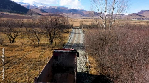 A red dump truck travels on a gravel road through a barren rural landscape with mountains in the background and clear winter skies.