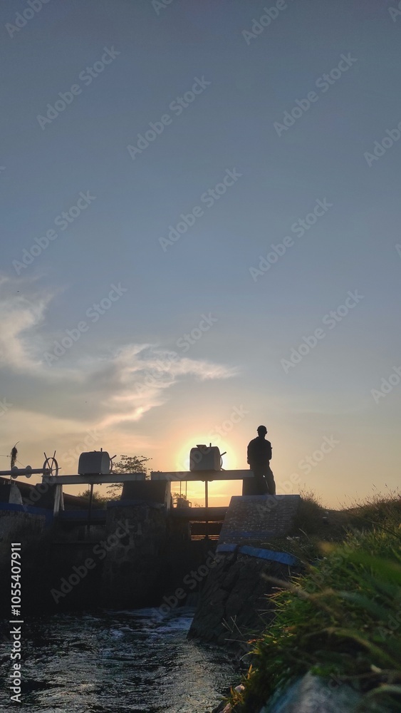 Fototapeta premium An unrecognised person stands amd taking picture on a water gate structure during sunset. The peaceful scene captures the calmness of dusk and the flow of water below