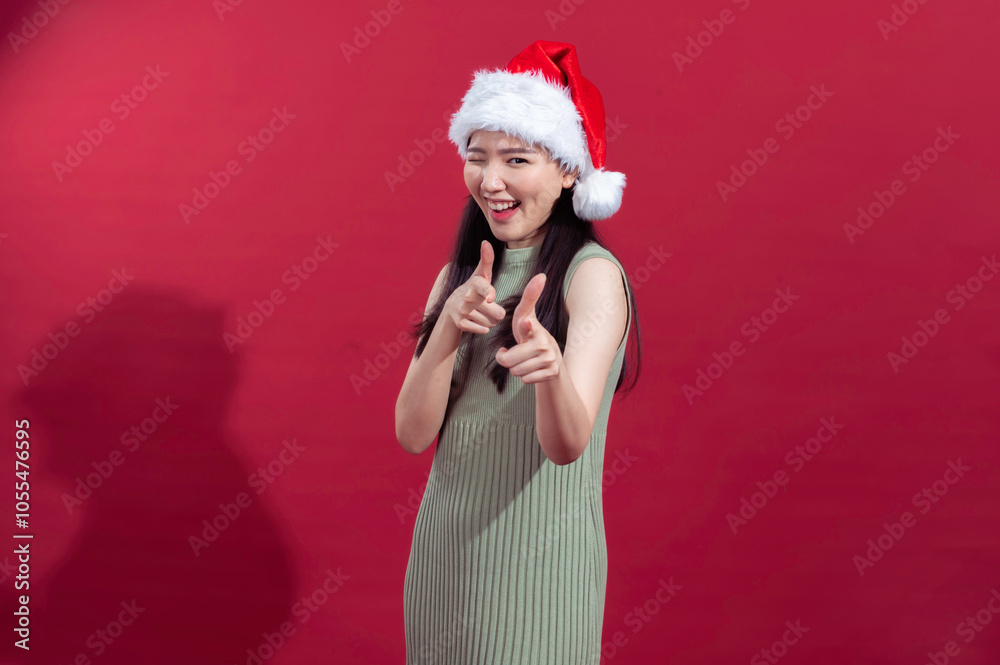 Asian woman wearing a Santa hat and green dress, standing against a red background. She winks and points both fingers towards the camera in a playful and energetic pose, expressing holiday cheer Asian woman wearing a Santa hat and green dress, standing against a red background. She winks and points both fingers towards the camera in a playful and energetic pose, expressing holiday cheer