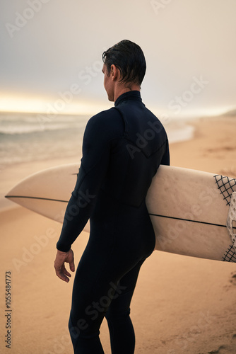 A surfer in a wetsuit holding a surfboard, gazing at the ocean from a sandy beach under an overcast sky.