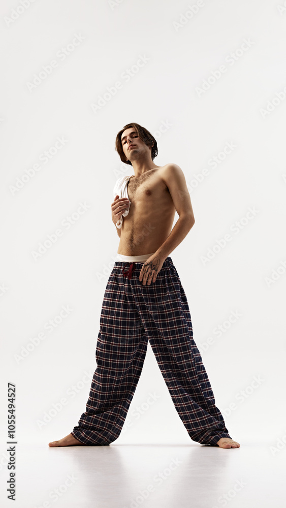 Fototapeta premium Man in plaid pants holding white shirt over his shoulder, looking up with confidence against white studio background. Concept of natural beauty, male health, fashion, skin care. Ad