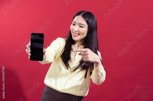 Asian woman smiling while extending a smartphone towards the camera, wearing a yellow sweater against a red background. The focus on the phone suggests a concept related photo