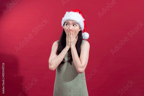 Asian woman wearing a Santa hat and green dress against a red background, with a surprised expression and hands covering her mouth, capturing a festive and playful reaction