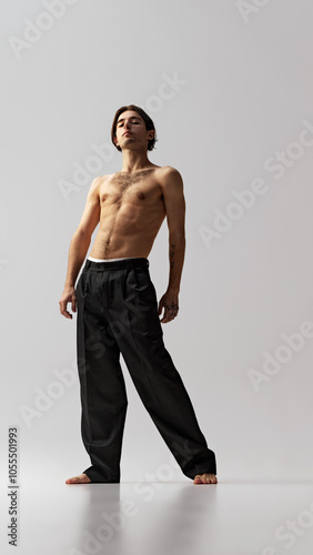 Full-length portrait of young athletic man in black pants posing showing independence and confidence against grey studio background. Concept of natural beauty, male health, fashion, skin care. Ad