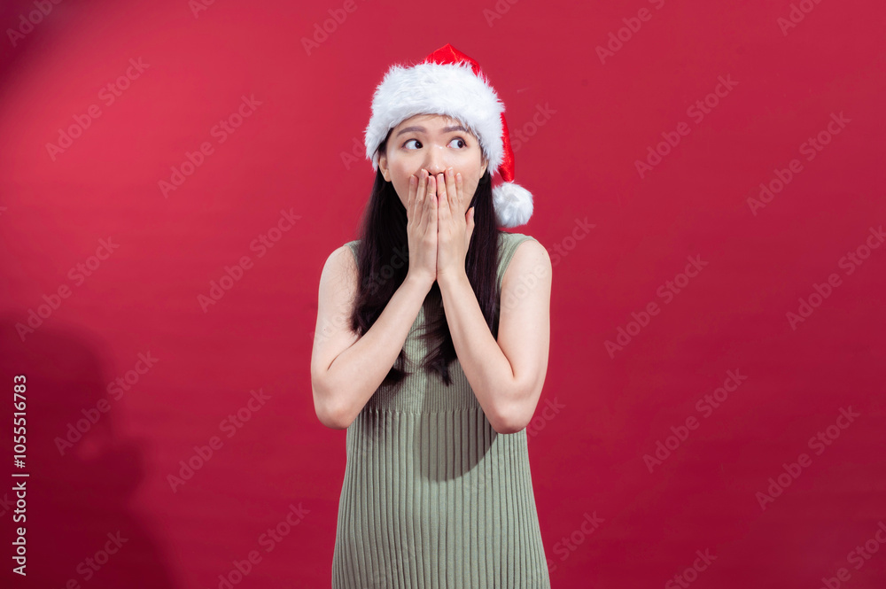 Asian woman wearing a Santa hat and green dress against a red background, with a surprised expression and hands covering her mouth, capturing a festive and playful reaction Asian woman wearing a Santa hat and green dress against a red background, with a surprised expression and hands covering her mouth, capturing a festive and playful reaction