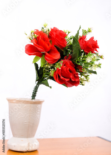 A bouquet of red flowers with green leaves and white filler flowers in a cream-colored vase with embossed designs, placed on a wooden surface against a white background.