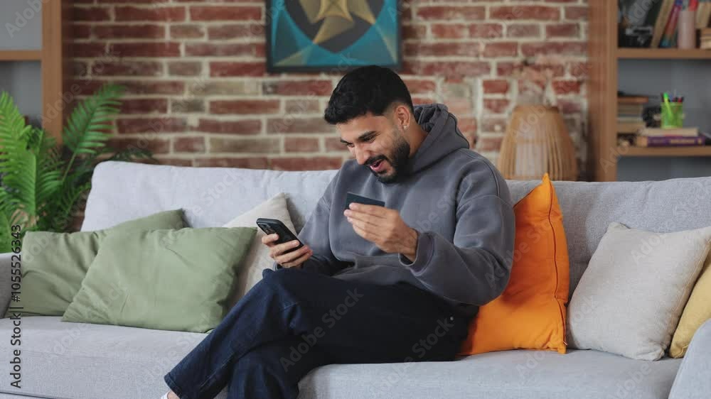 Amazed man in casual outfit screaming wow while keeping bank card and electronic device in hands. Young arabic person checking cyber wallet and noticing bonus crediting on screen of telephone. Amazed man in casual outfit screaming wow while keeping bank card and electronic device in hands. Young arabic person checking cyber wallet and noticing bonus crediting on screen of telephone.