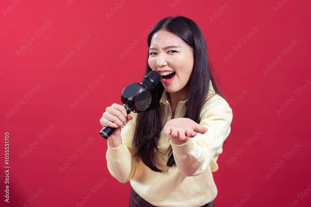 Obraz premium Asian woman smiling and singing into a microphone, wearing a yellow sweater with a red background. She appears enthusiastic and expressive, holding the microphone