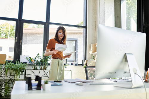 Фотография A young non binary individual stands at their desk, examining important papers in an airy workspace