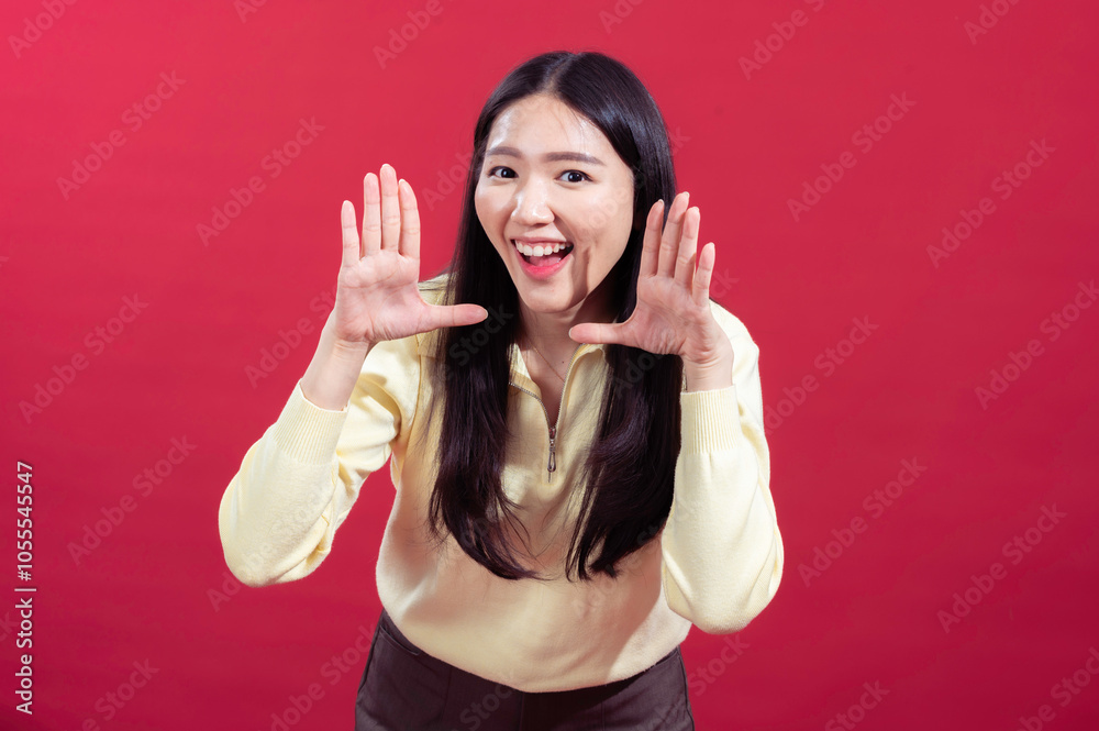 Asian woman posing playfully with hands framing her face, smiling widely, wearing a yellow sweater against a red background. Her expression is lively and engaging, showing enthusiasm and joy Asian woman posing playfully with hands framing her face, smiling widely, wearing a yellow sweater against a red background. Her expression is lively and engaging, showing enthusiasm and joy