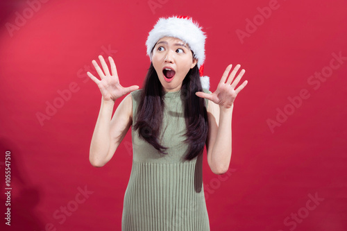 An Asian woman is joyfully raising both hands with an excited expression. She is wearing a Santa hat and a sleeveless green ribbed dress, standing in front of a solid red background photo