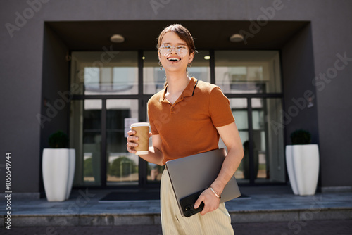 A non binary person stands joyfully outside their office, holding coffee and a laptop. photo