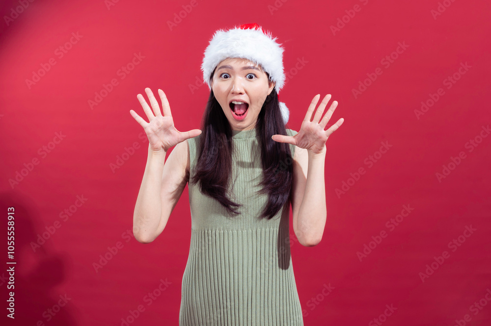 An Asian woman is joyfully raising both hands with an excited expression. She is wearing a Santa hat and a sleeveless green ribbed dress, standing in front of a solid red background An Asian woman is joyfully raising both hands with an excited expression. She is wearing a Santa hat and a sleeveless green ribbed dress, standing in front of a solid red background