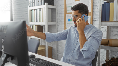 Young man with beard talking on smartphone in modern office interior, gesturing while looking at computer screen surrounded by shelves and office supplies