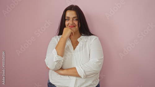 Young, hispanic, plus-size, woman smiling confidently against an isolated pink background, embodying body positivity and natural beauty in a casual white shirt and jeans