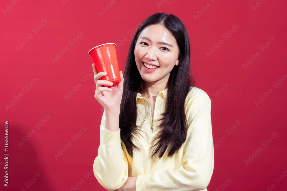 Asian woman smiling and holding a red plastic cup in her hand, wearing a yellow sweater against a red background. She looks cheerful and relaxed, with one arm crossed and the other holding cup Asian woman smiling and holding a red plastic cup in her hand, wearing a yellow sweater against a red background. She looks cheerful and relaxed, with one arm crossed and the other holding cup