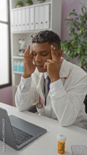 Young man in a white coat, stressed at a laptop in a clinic, with stethoscope, pill bottle, and office background.