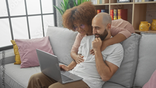 Interracial couple cuddling on a sofa while looking at a laptop in a cozy living room