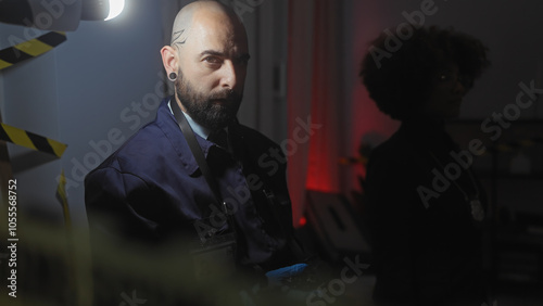 A man and a woman with detective badges investigate a dimly lit indoor crime scene with caution tape