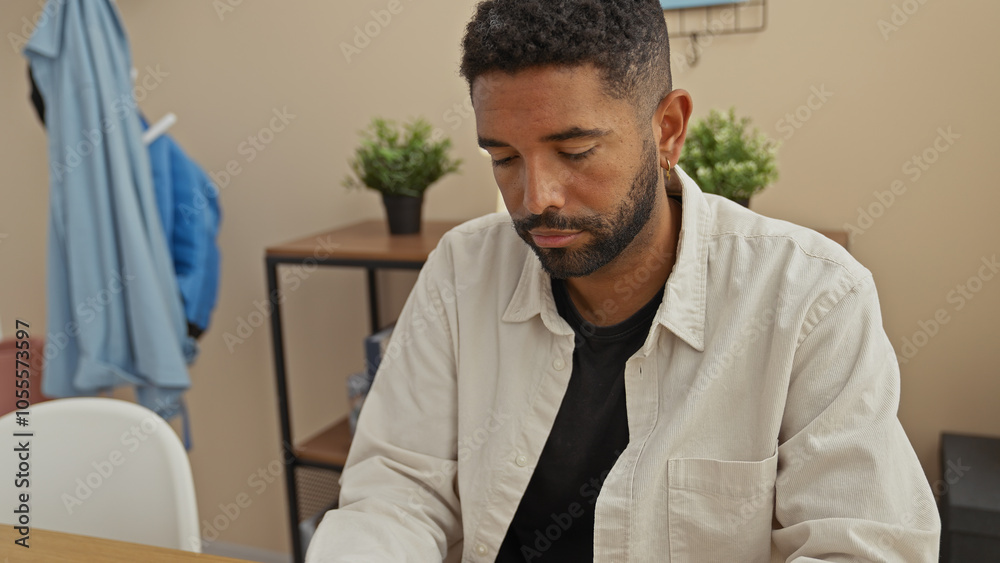 A contemplative young man with a beard sits indoors in a casual room, appearing thoughtful or fatigued. A contemplative young man with a beard sits indoors in a casual room, appearing thoughtful or fatigued.