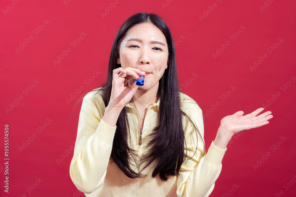 Asian woman in a yellow sweater blowing a blue party horn, with one hand raised in a playful gesture. She has a cheerful expression against a solid red background, creating a festive mood Asian woman in a yellow sweater blowing a blue party horn, with one hand raised in a playful gesture. She has a cheerful expression against a solid red background, creating a festive mood