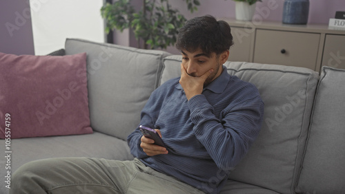 A thoughtful man with a beard sits on a sofa holding a smartphone in a modern living room.