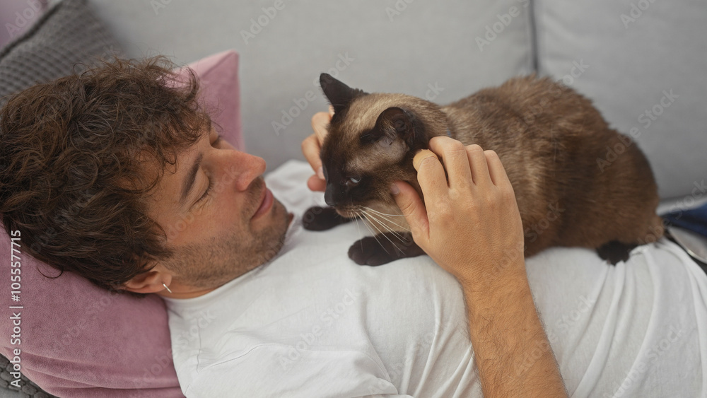 A young hispanic man lovingly pets his siamese cat while reclining on a sofa in a cozy living room. A young hispanic man lovingly pets his siamese cat while reclining on a sofa in a cozy living room.
